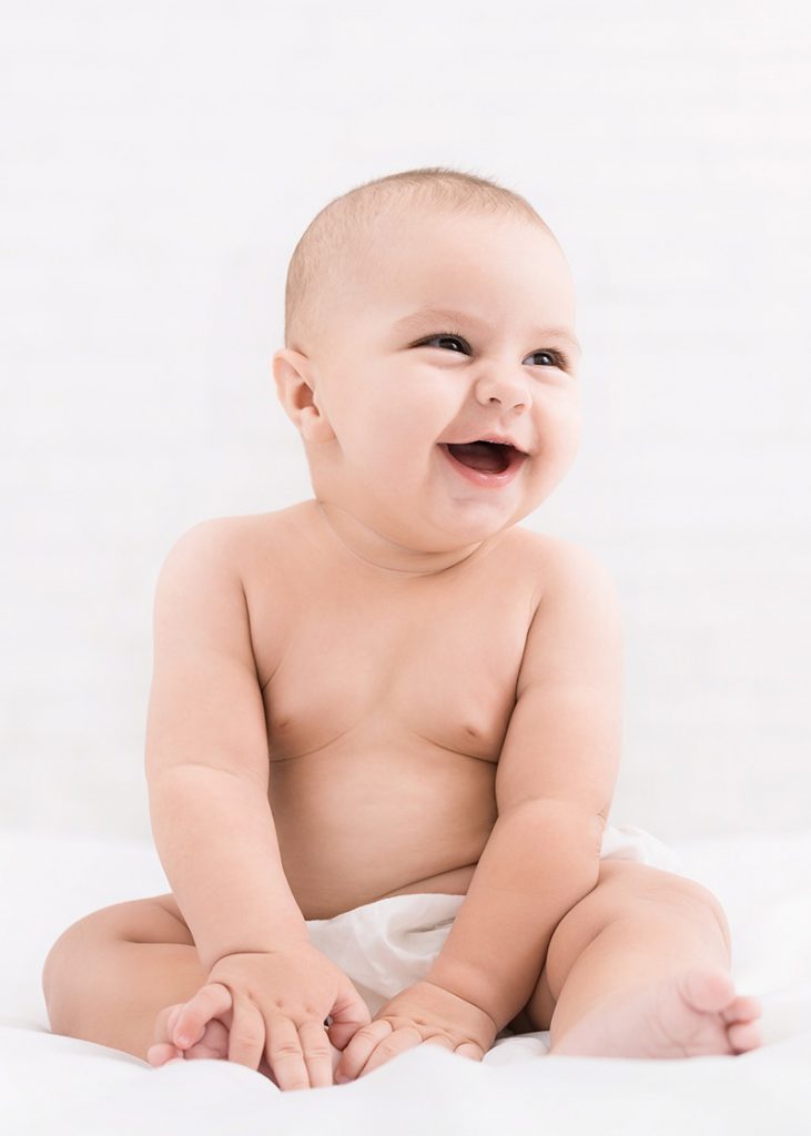 Adorable baby looking aside at copy space and smiling, sitting on bed on white background