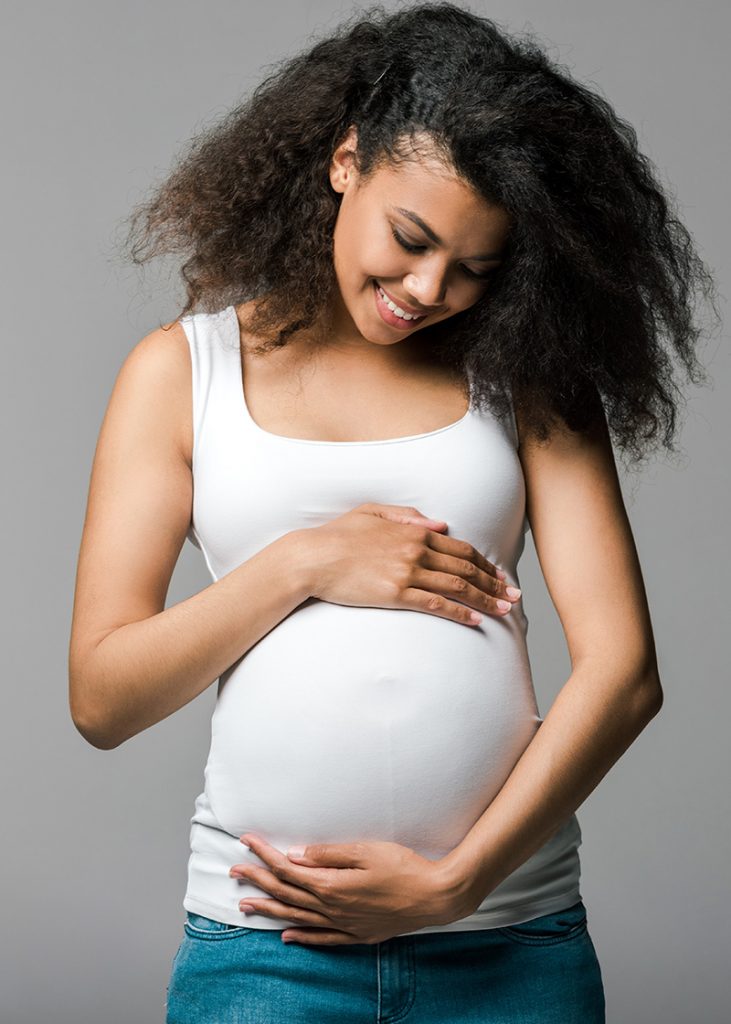Pregnant brown skinned woman holding rounded belly with gray background.